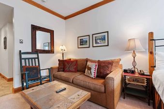 A cozy living area in The Charter D2205 features a brown sofa with decorative pillows, a tiled coffee table, a rocking chair, and framed artwork, creating a warm and inviting atmosphere.