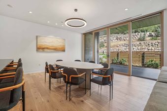 A modern dining room at Founders Place 2302 with an oval table and chairs, illuminated by contemporary lighting and offering views of a landscaped outdoor patio through large glass doors.