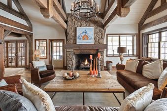 A cozy, rustic living room in Five Trees Estate with a stone fireplace, exposed wooden beams, and comfortable seating, bathed in natural light from large windows.