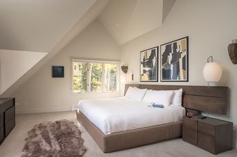 A serene and modern bedroom in Serenvale Retreat with a king-sized bed, contemporary furniture, abstract art, and a view of autumn trees through large windows.