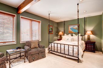 A serene bedroom featuring a four-poster bed with white bedding, two bedside tables with lamps, a cozy armchair, and two windows with wooden blinds.