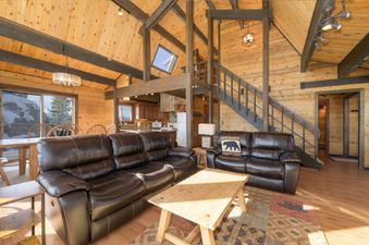The interior of 273 Fallen Rock is captured from the main living area, showcasing two brown leather sofas, a wooden coffee table, and a dining area, with a kitchen visible in the background and large windows revealing a snowy mountain exterior.