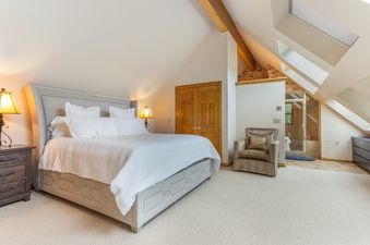 A cozy bedroom at Intrepid Retreat features a large bed with white linens, rustic nightstands, and unique angled ceilings with exposed wooden beams, creating a warm and inviting atmosphere.