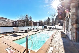 An outdoor hot tub at Lowell 301A steams under a bright blue sky, surrounded by lounge chairs, an outdoor dining set, and a scenic mountain backdrop.
