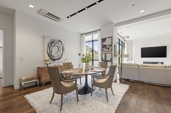 A modern dining area with a round wooden table, four chairs, and a white shag rug, illuminated by natural light and track lighting, with an open-concept lounge area in the background.