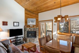 The Pines Condominiums A304's living and dining room with a stone fireplace and views of a snowy landscape.