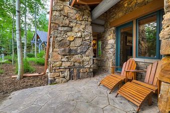 The private patio of Bear Paw Lodge 2 Bedroom features two wooden Adirondack chairs on a patterned stone surface, with a stone wall exterior and lush aspen trees in the background.