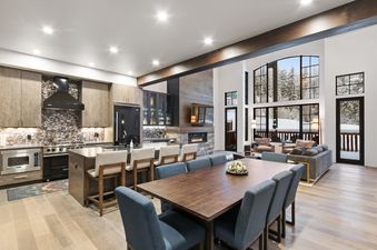 An expansive open-concept living space in Slopeside 129 features a modern kitchen, a large dining table, and a cozy living room with a fireplace and stunning views of a snow-covered forest through large windows.