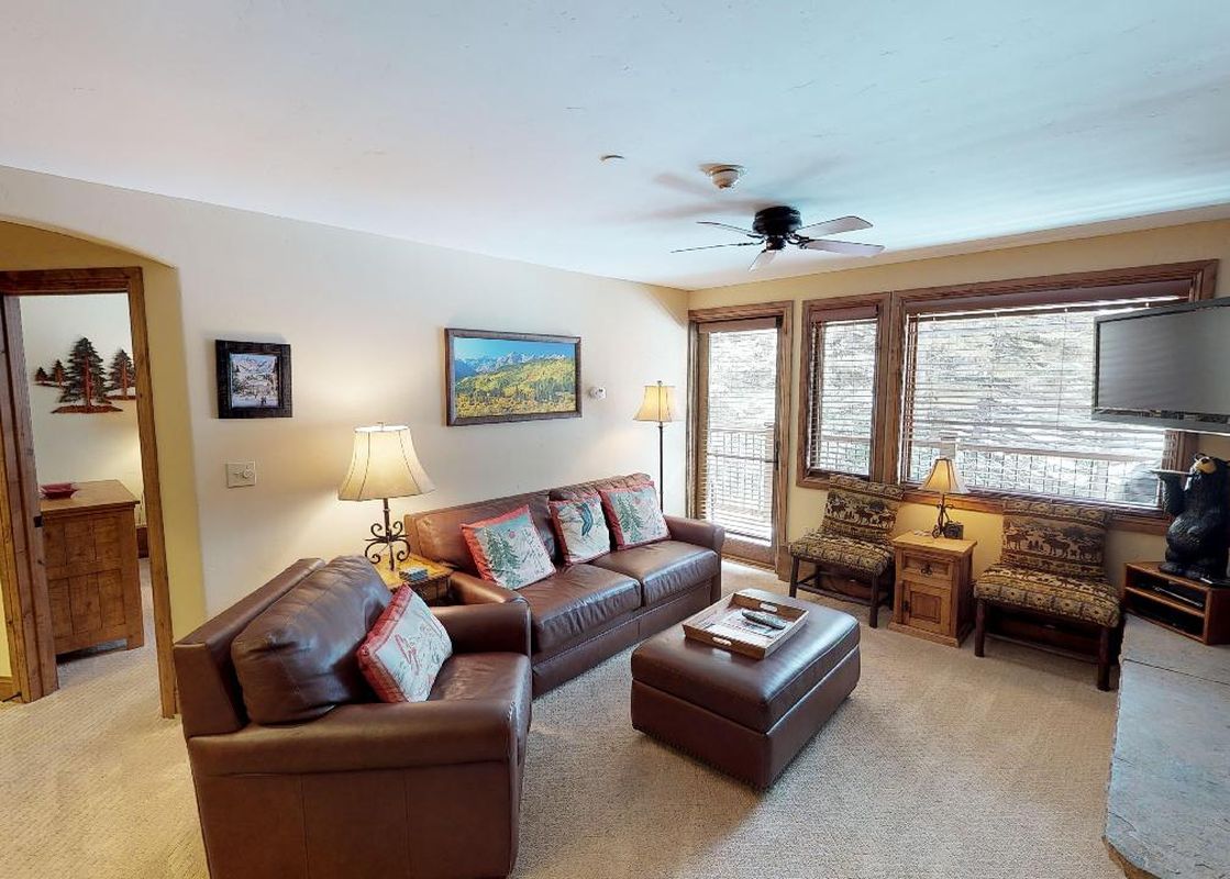 The living room of Manor Vail 267 features a brown leather couch, loveseat, and a coffee table. The room has a fireplace with a black bear statue on top and large windows with views of the snowy mountains outside.