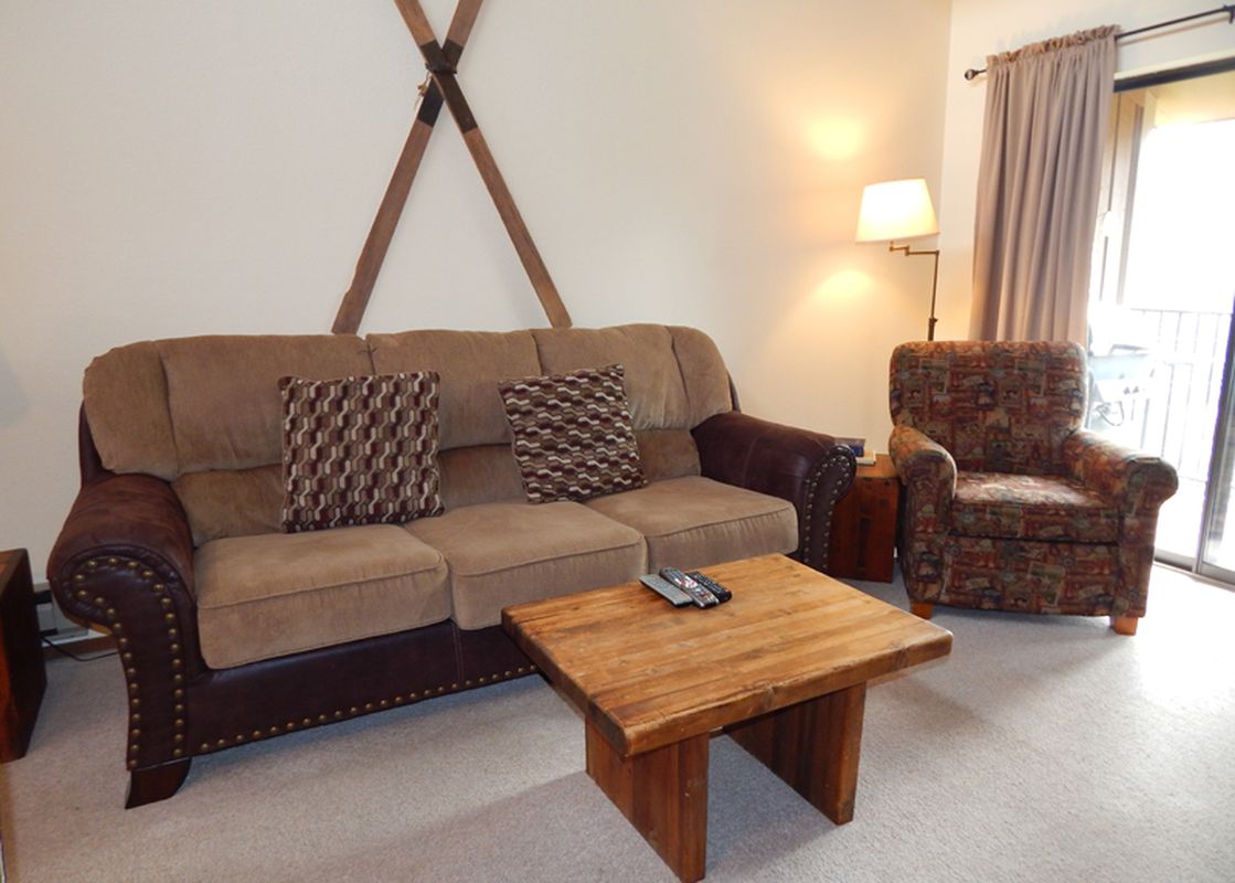 A cozy living room at Beaver Village 1932 features a comfortable sofa with decorative pillows, an armchair, a rustic wooden coffee table, and a floor lamp, with a large sliding glass door leading to the outside.