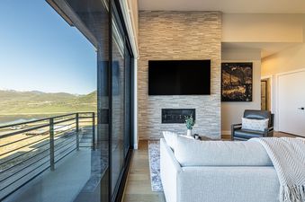 A modern living room with a large sliding glass door revealing a scenic mountain and lake view, a flat-screen TV above a fireplace, a light-colored sofa, and a dark leather armchair.