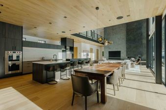 A modern home with a large kitchen and dining area with a wooden table and chairs. There are large windows that look out onto a forest.