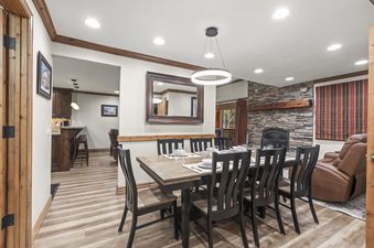 A modern dining area with a long wooden table, black chairs, and a contemporary circular chandelier, featuring a stone fireplace and a glimpse of the kitchen and living room.