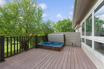 A private outdoor deck features a hot tub with its cover partially open, surrounded by a dark railing and lush green trees under a blue sky.