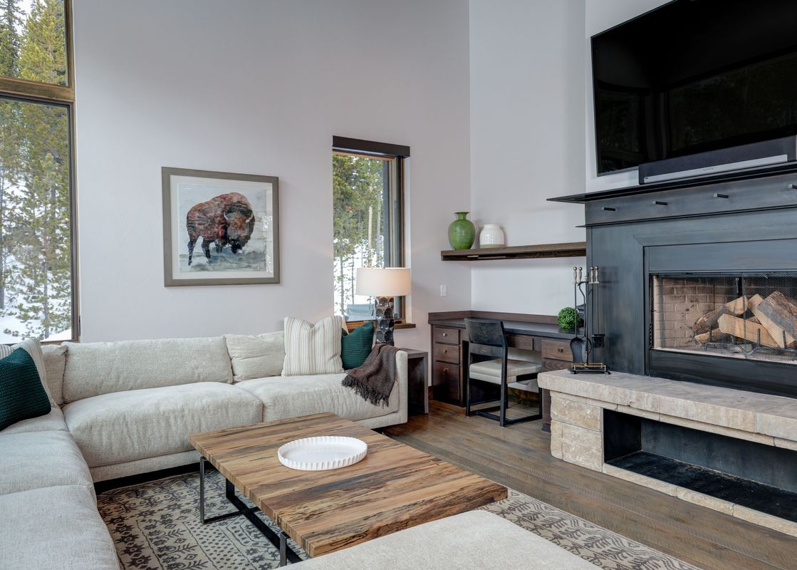 A spacious and elegantly furnished living room at Homestead Chalet 8 Claim Jumper, featuring a light-colored sectional sofa, a wooden coffee table, a fireplace, and large windows looking out onto a snowy pine forest.