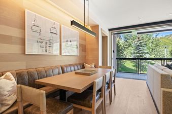 A modern dining area with a long wooden table, leather banquette seating, and chairs, featuring ski lift artwork on wooden paneled walls and an outdoor balcony view.