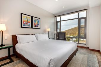 A luxurious king bedroom with a large window overlooking a scenic mountain landscape, featuring a comfortable bed, stylish nightstands, lamps, ski posters, and a dark leather armchair.