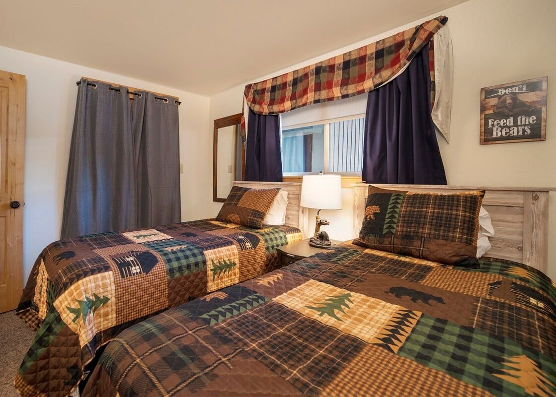 A rustic bedroom with two twin beds adorned with plaid and nature-themed patchwork quilts, a bedside table with a lamp, and a window with patterned curtains.