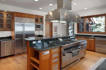 A spacious kitchen featuring stainless steel appliances, dark granite countertops, and warm wooden cabinetry, with a large window overlooking a snowy outdoor scene.