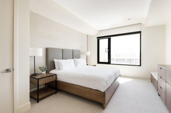 A bright, modern bedroom at Lift 406 features a king-size bed with a grey upholstered headboard, white linens, two dark wood nightstands with lamps, a large window offering natural light, and a chest of drawers.
