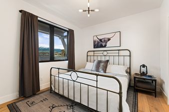 A cozy bedroom features a metal bed frame, white bedding, and a large window with mountain views, complemented by rustic decor and a highland cow artwork.