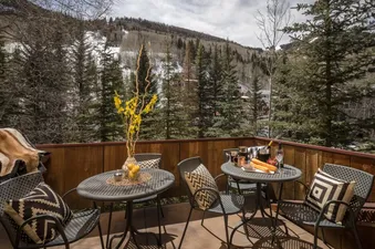 The private balcony at River Bliss features two bistro tables and chairs with patterned pillows, set against a backdrop of evergreen trees and snowy mountains.