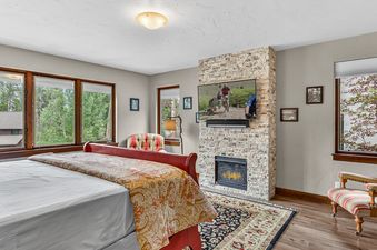 A cozy bedroom featuring a fireplace, a king-sized bed with a red frame, and large windows overlooking lush greenery.
