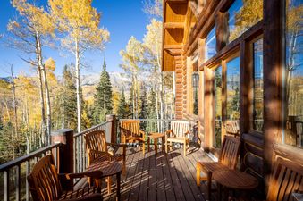 A wooden deck with chairs and tables overlooks golden aspen trees and distant mountains, set against a clear blue sky, showcasing the exterior of Bear Lodge.