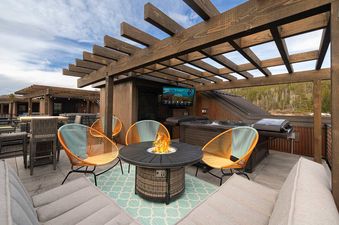 A modern outdoor deck with a fire pit, stylish chairs, and a hot tub, set under a wooden pergola with a view of a forest.