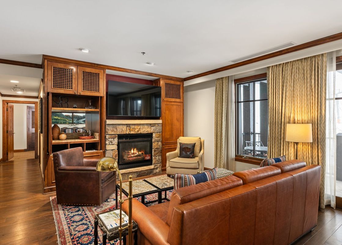 A warm and inviting living room at Elkhorn Premier 8405 features a stone fireplace, integrated TV, and cozy leather seating, complemented by an outdoor balcony view.