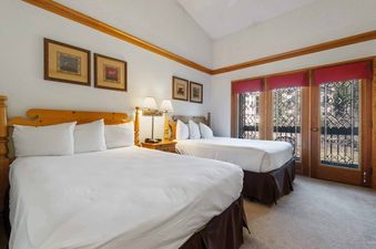 A cozy bedroom at The Charter E2105 features two queen beds with white linens, wooden headboards, a shared nightstand with a lamp, and windows with red valances overlooking the exterior.