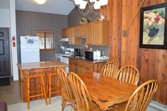 The image shows the kitchen and dining area of Beaver Village 1633, featuring a rustic decor with wooden cabinets, a granite-topped island, and a dining table with chairs.