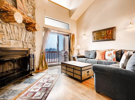 A cozy living room at Powder Pointe 303B with a stone fireplace, gray sofas, a trunk coffee table, and a balcony view of a snowy outdoor scene.