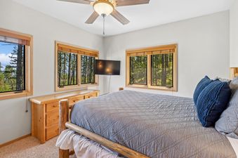 A cozy bedroom at Wood Run Ski Haus features a rustic wooden bed frame with a blue quilted comforter, a wall-mounted TV, and large windows overlooking a dense forest.