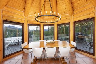 A rustic dining room with a large wooden table and white chairs, surrounded by windows offering mountain and forest views, under a grand circular chandelier.