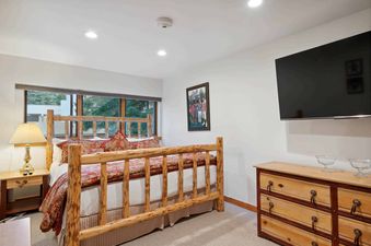A cozy bedroom with a rustic log bed, vibrant red and gold bedding, a bedside table with a lamp, a large window offering a view of greenery, and a wooden dresser with a wall-mounted television.