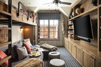 A rustic bunk room featuring wooden bunk beds, a sofa with pillows, a flat-screen TV, built-in shelves, and a window with a blue patterned shade.