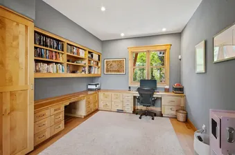A home office with custom wooden desks, extensive bookshelves filled with books, an ergonomic office chair, and a large window overlooking green trees.