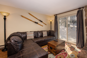 A spacious living area featuring a large brown sectional sofa, a wooden coffee table, and a colorful patterned rug. The room is illuminated by two floor lamps, and vintage wooden skis adorn one wall, while a glass sliding door reveals a snowy outdoor scen