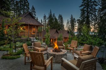 A cozy outdoor seating area with a lit fire pit surrounded by comfortable chairs at Cedar Crest Sparrow Cottage 1, set against a backdrop of cabins and pine trees at dusk.