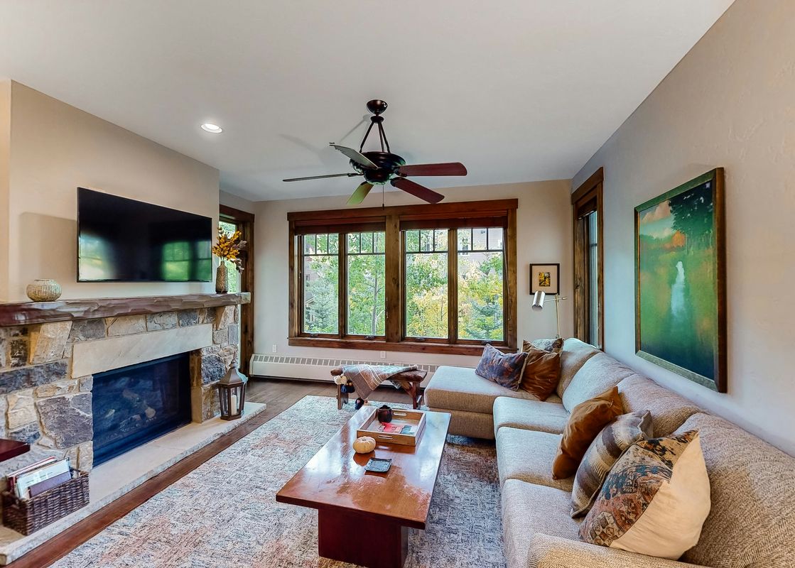 A spacious and inviting living room at Water House On Main Street 5205 with a large sectional sofa, stone fireplace, and views of lush greenery through large windows.