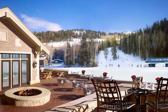 A large stone patio with a lit fire pit and outdoor seating overlooks a snowy mountain ski slope with pine trees and skiers.