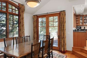 The dining area at Jay Chalet features a large wooden table and chairs, with windows offering views of snow-covered trees, and an adjacent kitchen with wooden cabinets and black countertops.