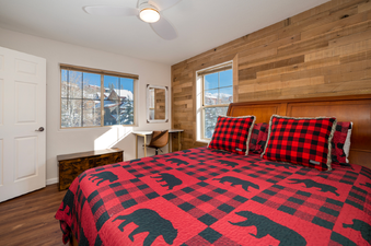 A cozy bedroom with a red and black plaid bedspread featuring bear silhouettes, two windows showcasing a snowy winter landscape, and a light wood-paneled accent wall.