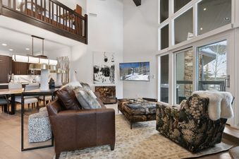 The spacious living area features a brown leather sofa and a patterned armchair, with a large cowhide ottoman in the center, and a modern television and ski-themed artwork on the wall, and high windows providing natural light.