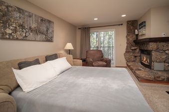 A cozy living room in Beaver Village 1624 features a pull-out sofa bed with light blue bedding, a stone fireplace, and a glass door leading to a balcony overlooking a pine forest.