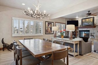 The dining area of The Charter C2205 features a large wooden table and chairs under an antler chandelier, with a console table leading to the living room with a fireplace.