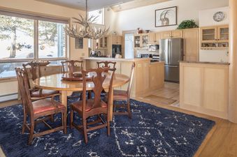 A warm and inviting dining area and kitchen at Hillside Hideaway, featuring a large wooden dining table, antler chandelier, and expansive windows with a snowy view.