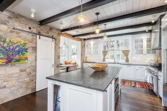 A modern kitchen with white cabinetry, a large island with a dark countertop, and rustic lighting fixtures. There is a stone accent wall and a breakfast nook.