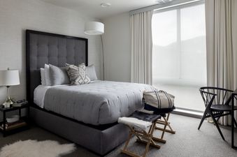 A serene bedroom in Penthouse on Glenwood 401 featuring a grey tufted bed, a modern nightstand with a lamp, a large window with white curtains, and two wooden folding chairs.